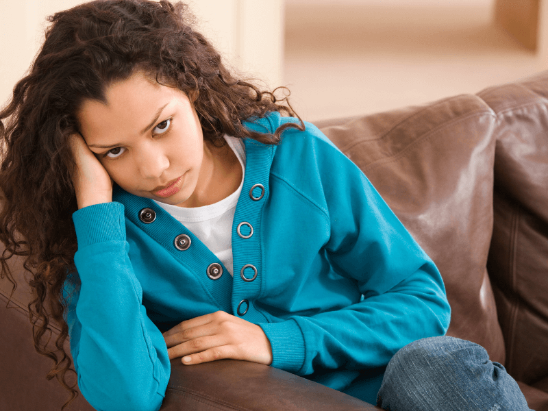 Adolescent black girl looking sad and leaning her head on her hand while sitting on a couch