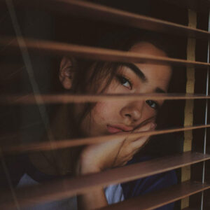young teen girl looking out the window that has blinds as she struggles with various mental health conditions