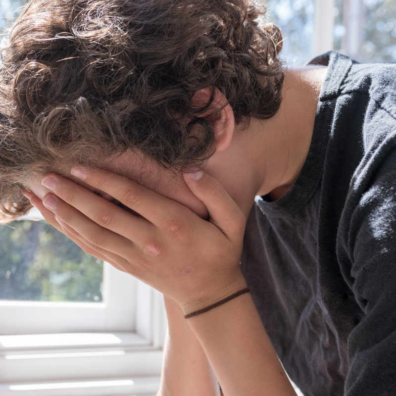 Adolescent boy sitting next to a window with his hands covering his face struggling with mental health