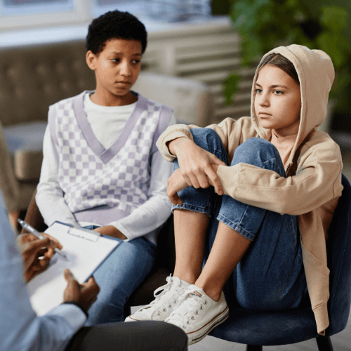 Am early-teen girl has her feet in a chair as a girl next to her looks at her. Their partially seen therapist sits across from them.