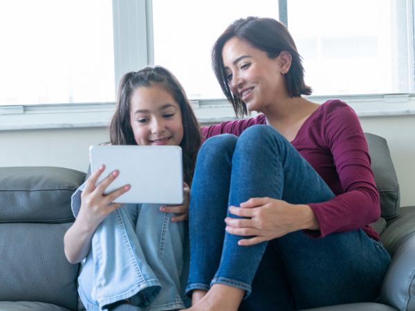 An adolescent girl and her mother sitting on the couch while using a tablet for virtual therapy
