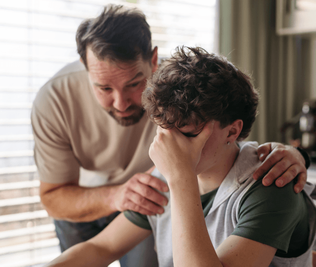 A struggling teen is sitting at the kitchen table as his father has his hands on the boy's shoulders