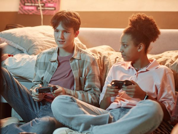 A teen girl looks at a teen boy while they sit on the floor at home holding video game controllers while playing