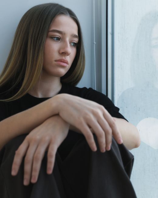 A sad teen girl sitting on a windowsill and looking outdoors