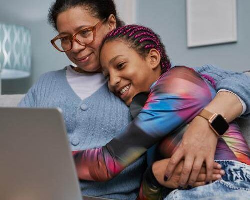 A mother and teen daughter sit on a couch, hugging during online therapy