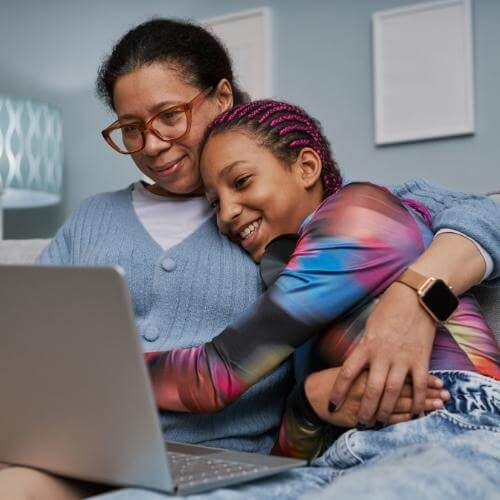 A mother and teen daughter sit on a couch, hugging during online therapy