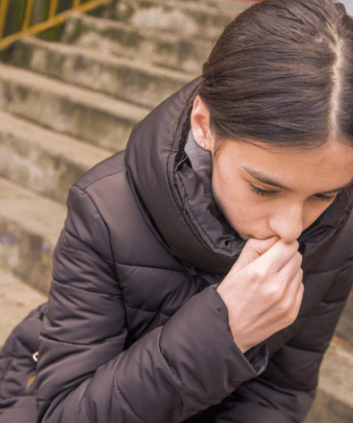 Young teen girl crouching outdoors on the steps with her head down and her hand to her mouth