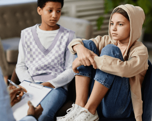 Am early-teen girl has her feet in a chair as a girl next to her looks at her. Their partially seen therapist sits across from them.