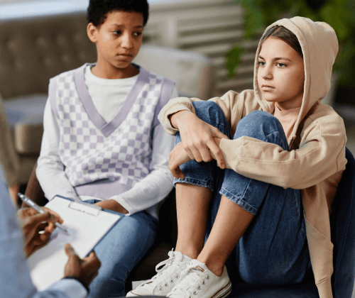 Am early-teen girl has her feet in a chair as a girl next to her looks at her. Their partially seen therapist sits across from them.