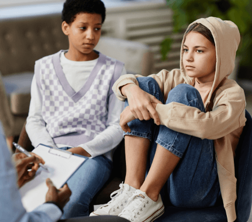 Am early-teen girl has her feet in a chair as a girl next to her looks at her. Their partially seen therapist sits across from them.