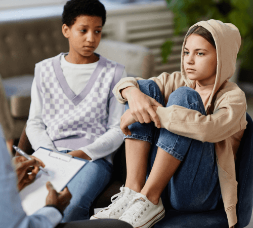 Am early-teen girl has her feet in a chair as a girl next to her looks at her. Their partially seen therapist sits across from them.