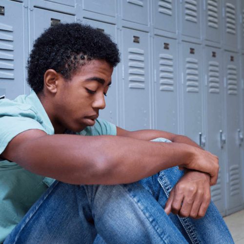 African American adolescent boy sitting in front of school lockers with his head down