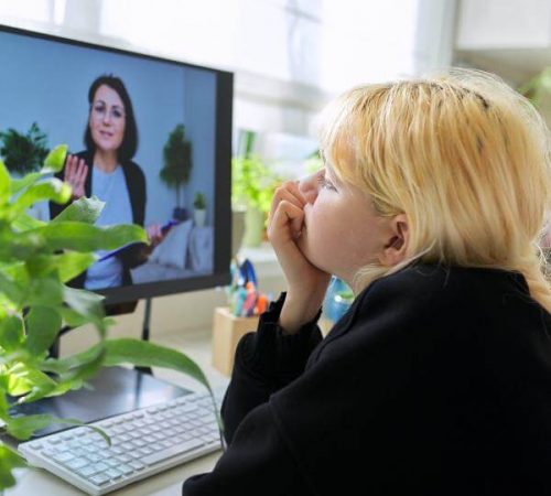 An adolescent girl sits at a desk with a computer and listens to her online therapist