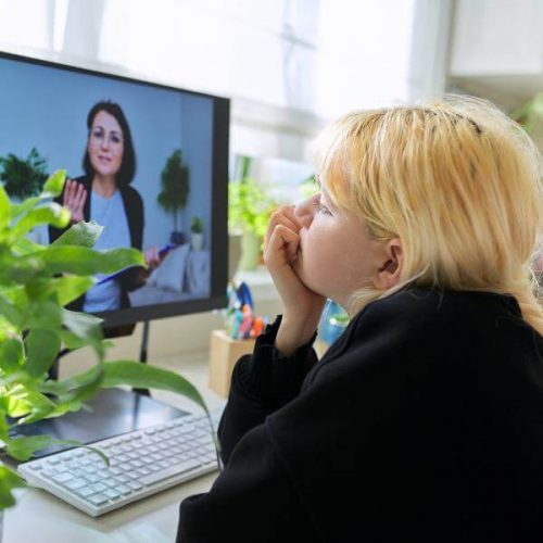 An adolescent girl sits at a desk with a computer and listens to her online therapist