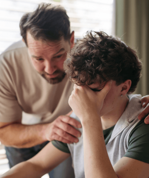 A struggling teen is sitting at the kitchen table as his father has his hands on the boy's shoulders