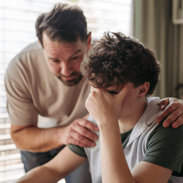 A struggling teen is sitting at the kitchen table as his father has his hands on the boy's shoulders