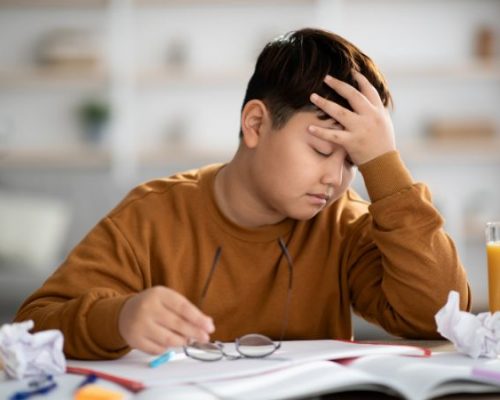 An adolescent boy is exhausted as he sits at a table trying to do homework