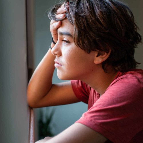 Adolescent boy with dark hair looking leaning on a window sill and sadly looking out the window