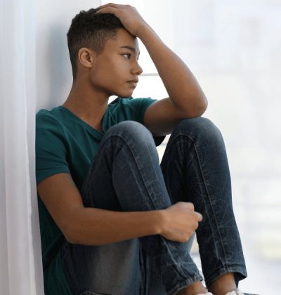 Teenage boy with anxiety siting indoors on a window ledge and looking outside