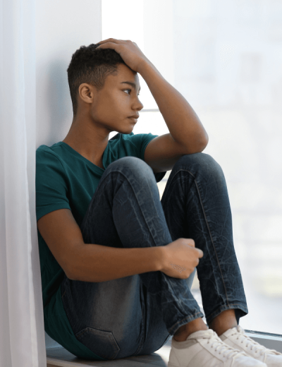 Teenage boy with anxiety siting indoors on a window ledge and looking outside