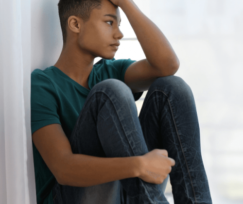 Teenage boy with anxiety siting indoors on a window ledge and looking outside