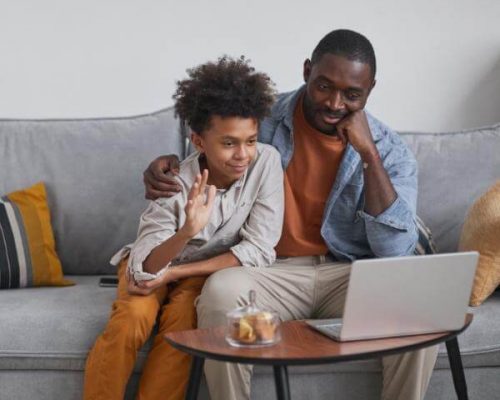 A father and son sit on a couch as the son greets his virtual therapist