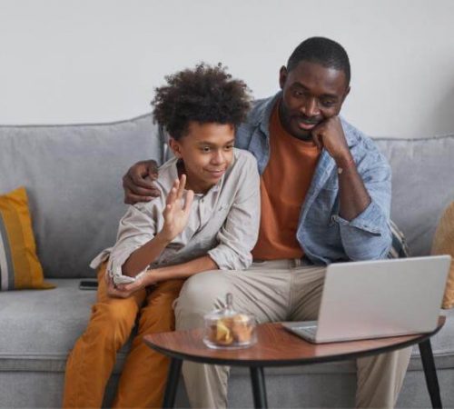 A father and son sit on a couch as the son greets his virtual therapist