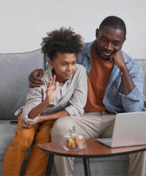 A father and son sit on a couch as the son greets his virtual therapist