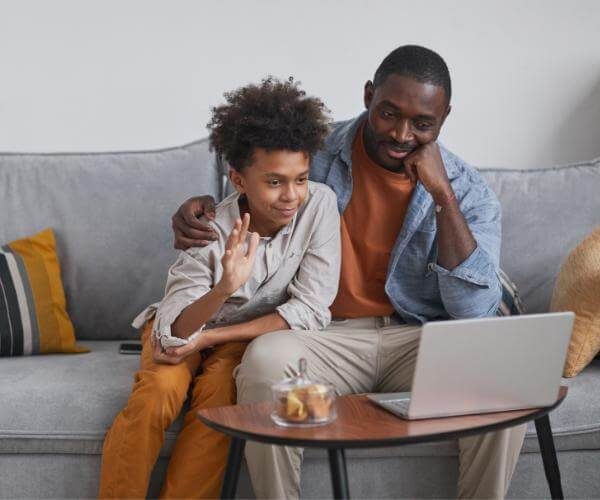 A father and son sit on a couch as the son greets his virtual therapist