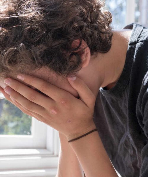 Adolescent boy sitting next to a window with his hands covering his face struggling with mental health